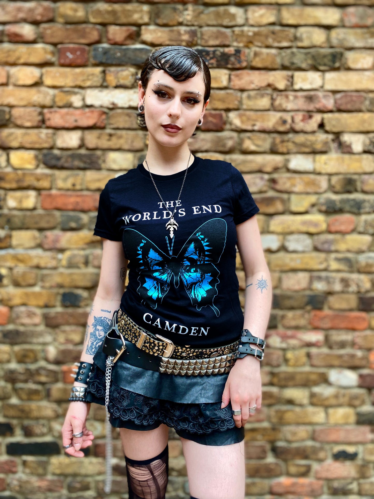 Person wearing a black t-shirt with a butterfly design and text, standing against a brick wall.
