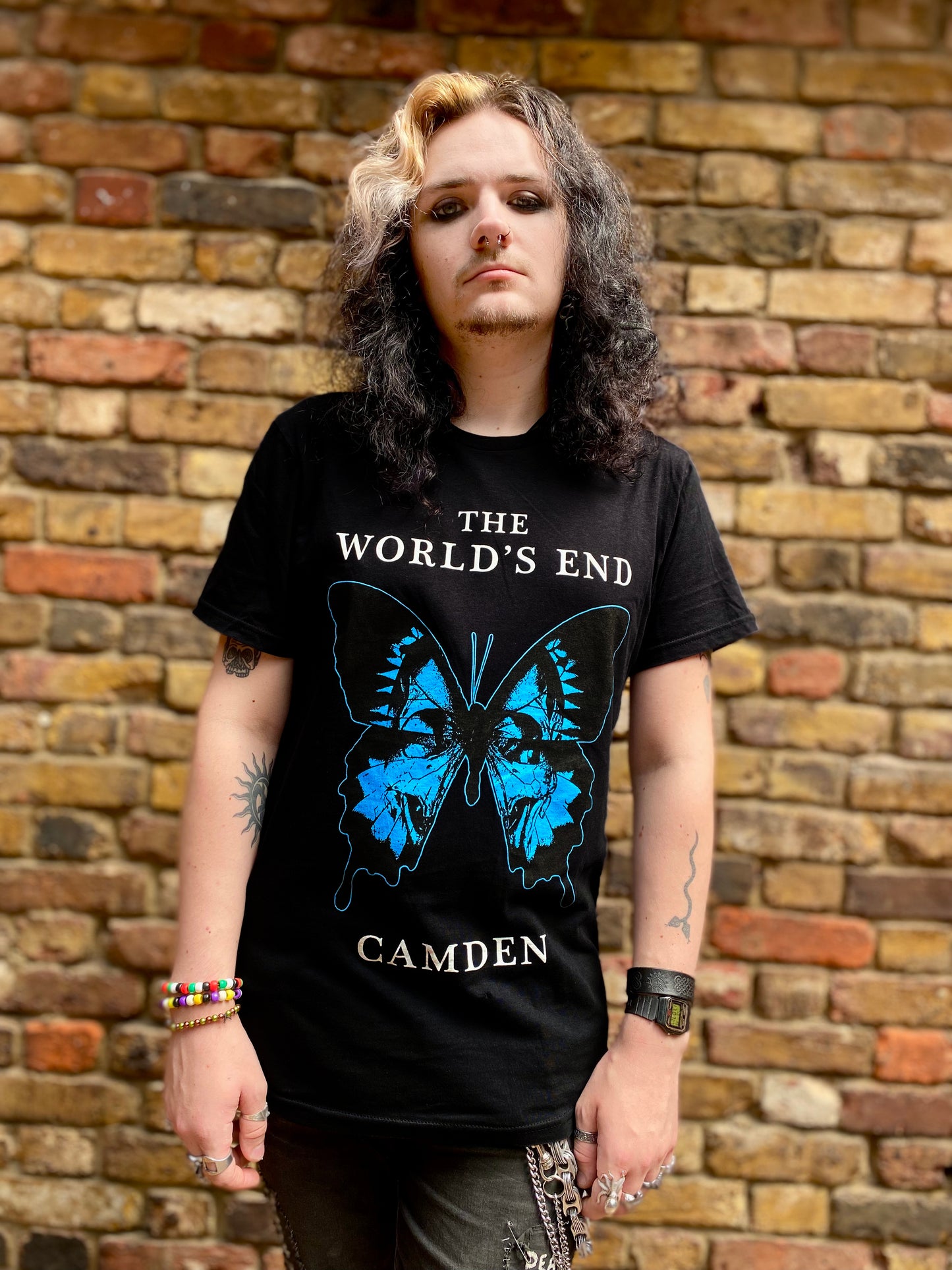 Person wearing a black t-shirt with a blue butterfly design and text, standing against a brick wall.
